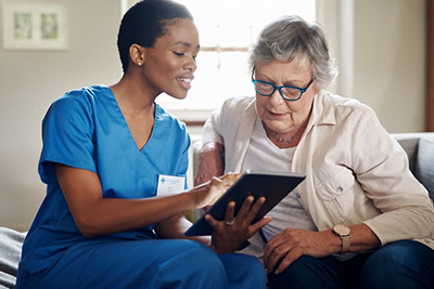 Young female home health nurse sitting with an elderly patient and looking at at an iPad screen together smiling