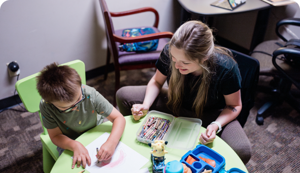 Image of a young female healthcare worker in business casual clothes who is working alongside a young child. They are sitting at a table and the child is coloring with crayons