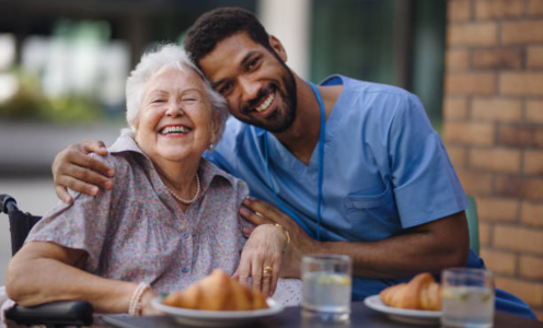 Image of a young male healthcare worker outside eating lunch with an elderly female woman who is smiling. He has his arm wrapped around her and they are both smiling for a posed picture. The table has croissants and glasses of water on it