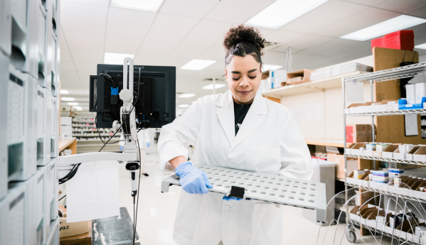 Person in a lab coat working in a pharmacy or lab, holding a tray.