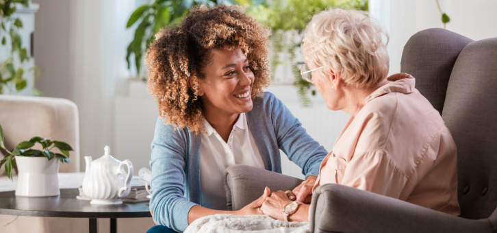 A young woman and an elderly woman smiling at each other in a cozy living room.
