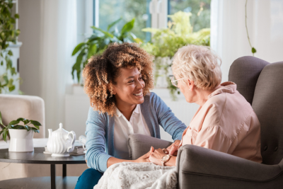 A young woman holds hands with an older woman in a sunlit living room.