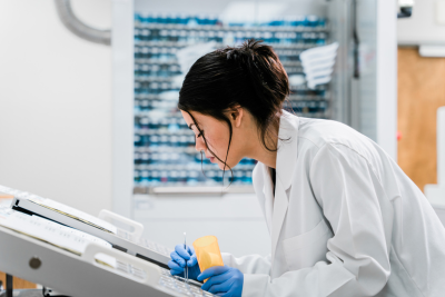 A woman in a lab coat and gloves works at a laboratory bench.