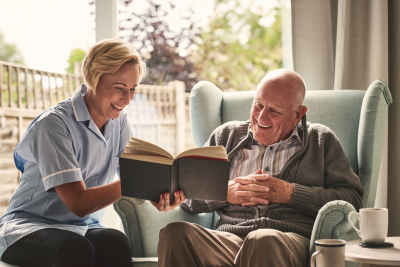 Image of a smiling female caregiver who is sitting in a comfortable home setting and reading a book to an elderly smiling male senior. They are also enjoying coffee that is sitting on the side table