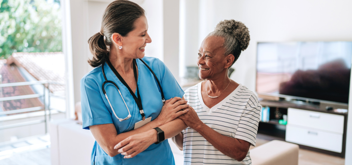 Image of a young female nurse at an elderly female patients home, they are both smiling and their arms are wrapped in one another as they communicate