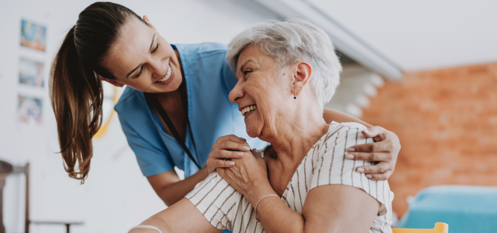 A caregiver in blue scrubs smiles at an older adult sitting down, both appear happy.