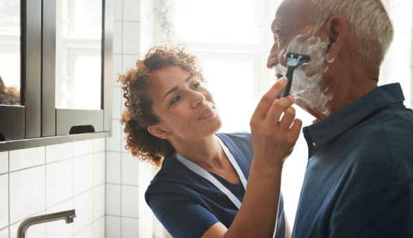 A woman in blue scrubs helps an elderly man shave, with shaving cream on his face, in a bright bathroom.