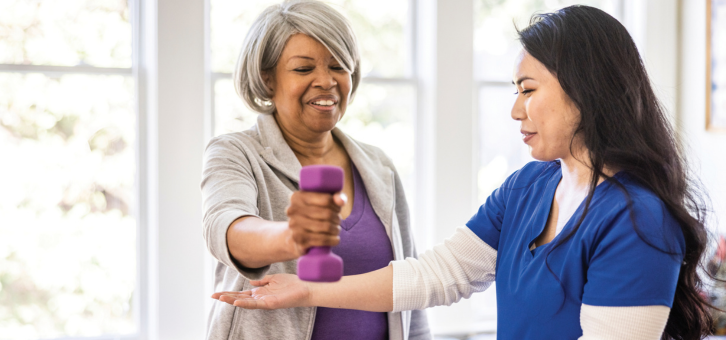 An older woman holds a purple dumbbell while a younger woman assists her with the exercise.