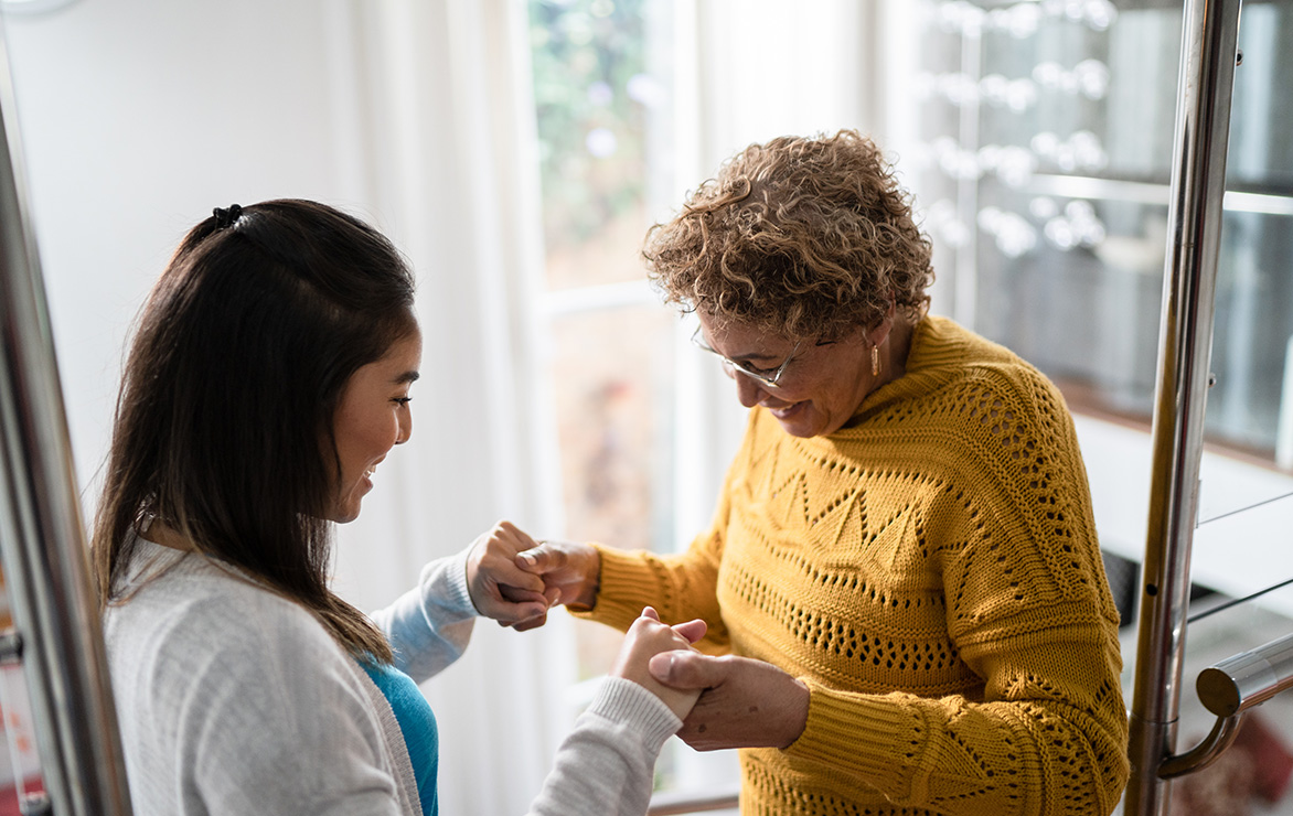 Two women smiling and holding hands in a well-lit room near a staircase.