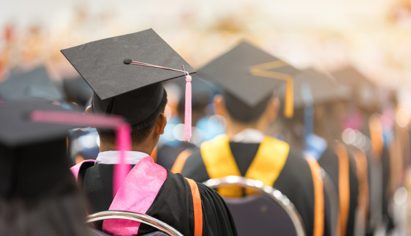 Graduates seated in a hall wearing caps and gowns, viewed from behind.