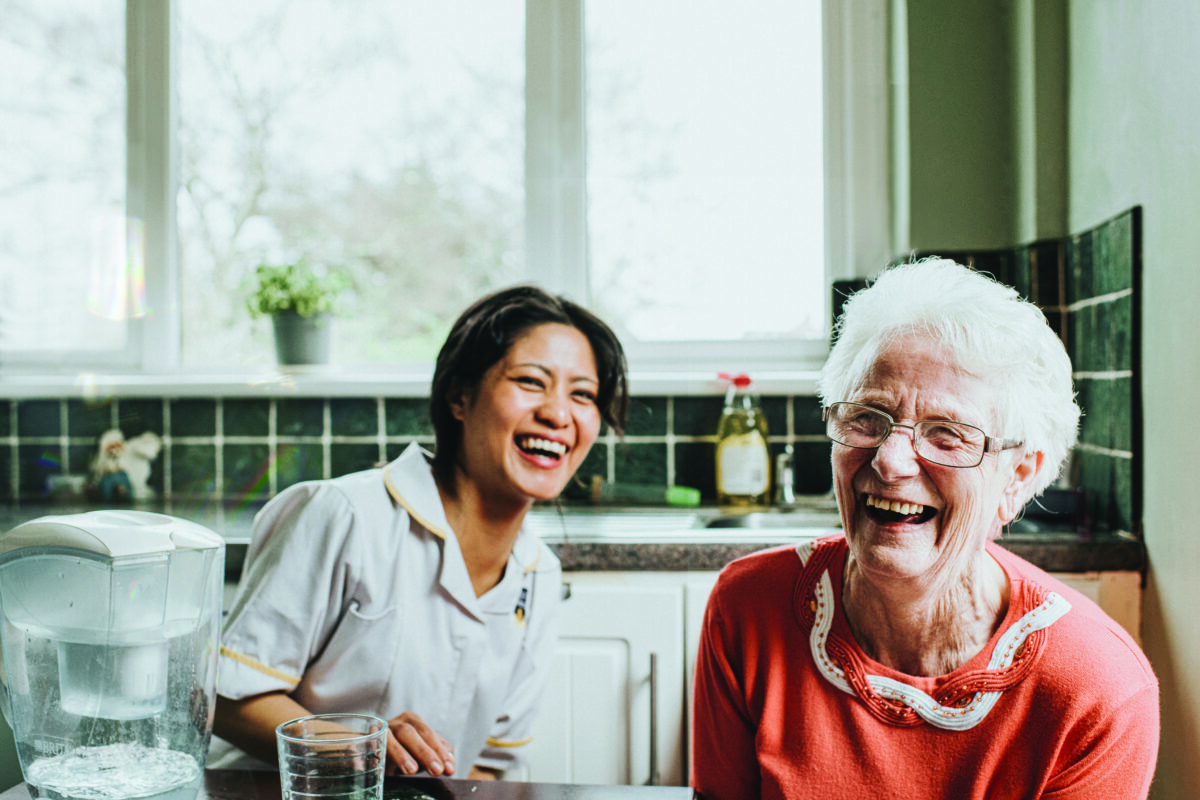 A young, friendly and bubbly caretaker sits beside an elderly woman at a kitchen table. The older woman faces the camera directly and laughs as the younger woman laughs behind her. The scene is lighthearted and fun, and the older woman quite clearly has a big personality.