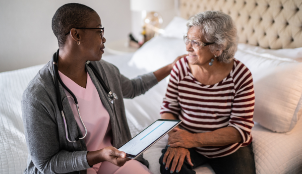 A healthcare professional talks to an elderly person sitting on a bed.