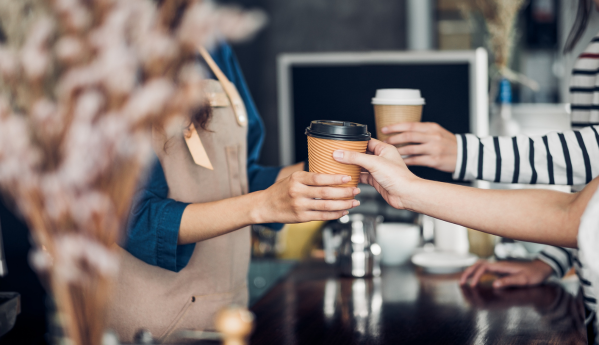 A barista hands a coffee to a customer over a counter in a coffee shop. Another person holds a cup in the background.