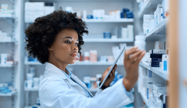 A pharmacist holding a tablet, reaching for a medication bottle on a pharmacy shelf.