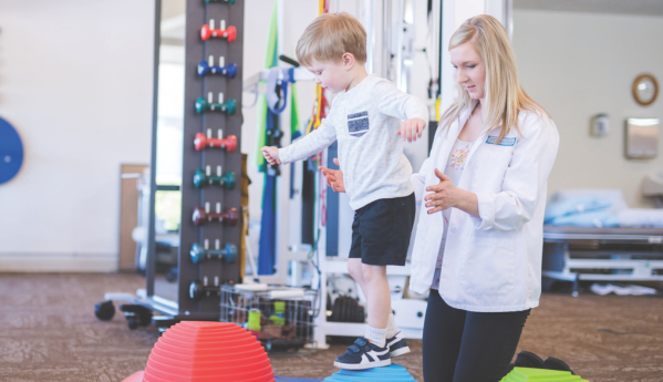 A young boy balancing on colorful platforms with a therapist's guidance in a gym setting.